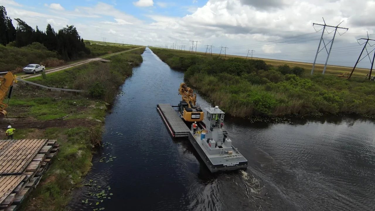 Tug and drill barge moving through a marsh channel.